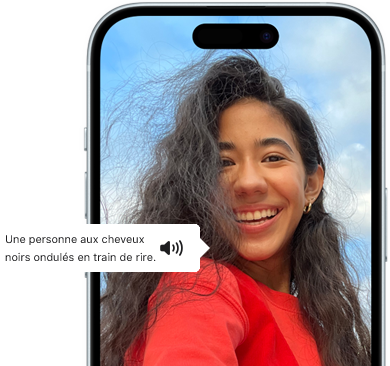 Un iPhone 15 montrant la fonctionnalité VoiceOver énonçant les informations figurant sur l’image : Une personne aux cheveux noirs ondulés en train de rire