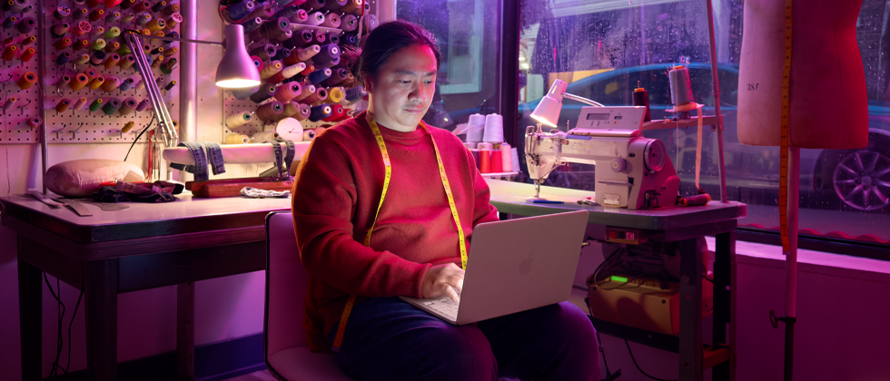 Une personne utilise un MacBook Neo, rose poudré, débranché et posé sur ses genoux, dans un espace de travail avec une machine à coudre et du matériel de couture, notamment du fil et un mannequin de couture