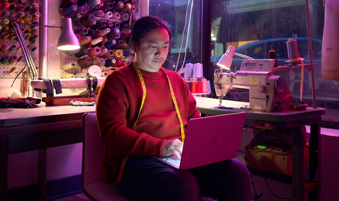 Une personne utilise un MacBook Neo, rose poudré, débranché et posé sur ses genoux, dans un espace de travail avec une machine à coudre et du matériel de couture, notamment du fil et un mannequin de couture