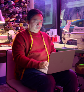 Une personne utilise un MacBook Neo, rose poudré, débranché et posé sur ses genoux, dans un espace de travail avec une machine à coudre et du matériel de couture, notamment du fil et un mannequin de couture